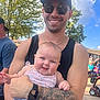 man, baby, smile, outdoor, sunglasses, tank_top, tattoo, arm, child, holding, happy, people, trees, daylight, casual_clothing, denim, floral, background, scooter, sunny