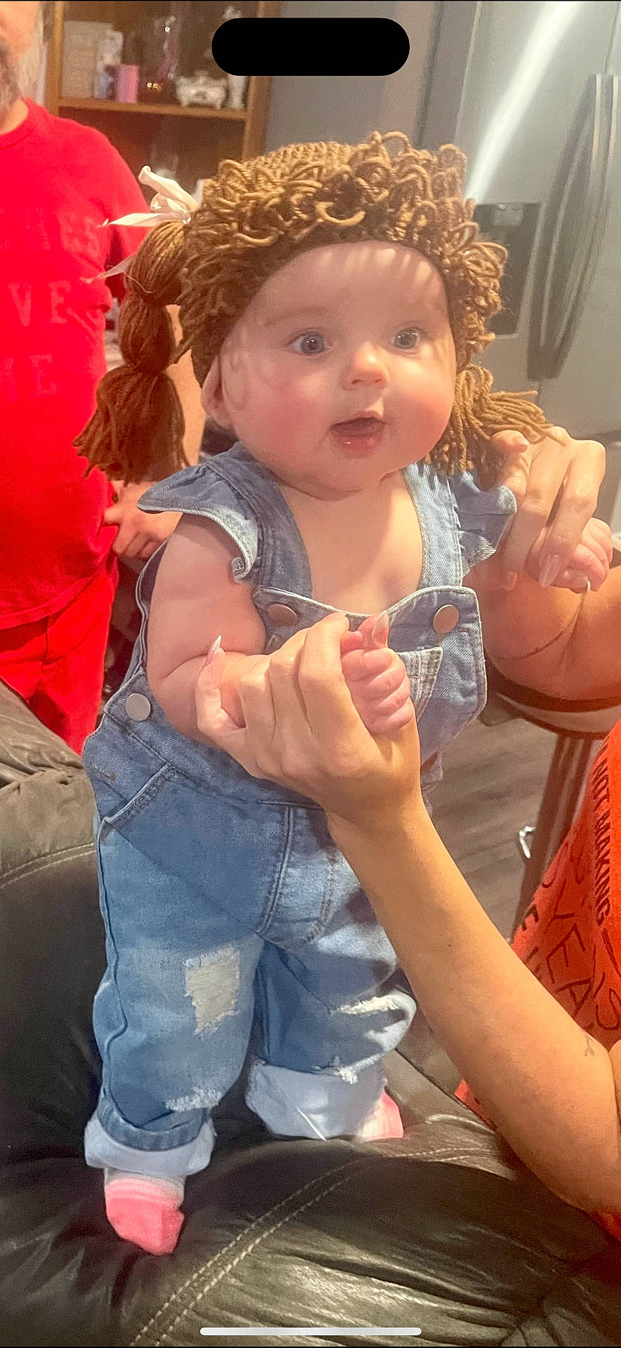 adult, baby, casual_clothing, child, couch, curious, cute, denim_overalls, furniture, hand_holding, home, indoor, living_room, person, pigtails, pink_socks, portrait, smiling, wide_eyes, yarn_wig