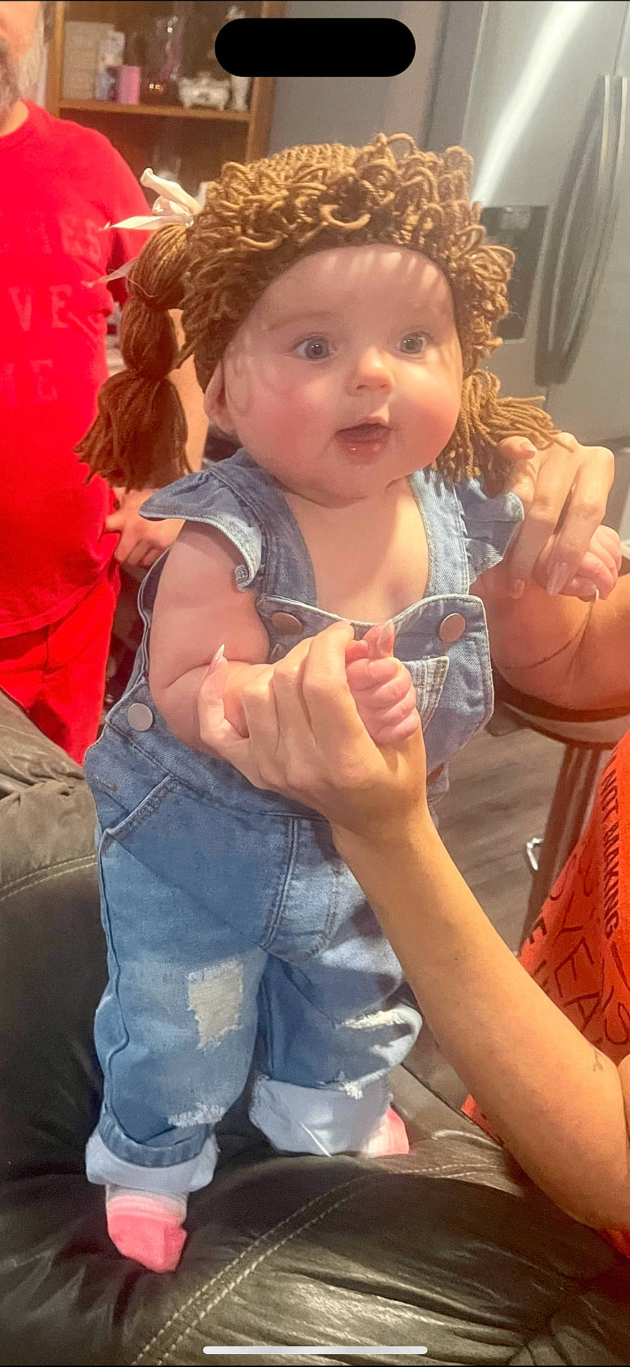 adult_hand, baby, casual_clothing, child, couch, curious_expression, denim_overalls, face, hand_holding, home_interior, indoor, kitchen, light, person, pigtails, pink_socks, refrigerator, smile, soft_light, yarn_wig