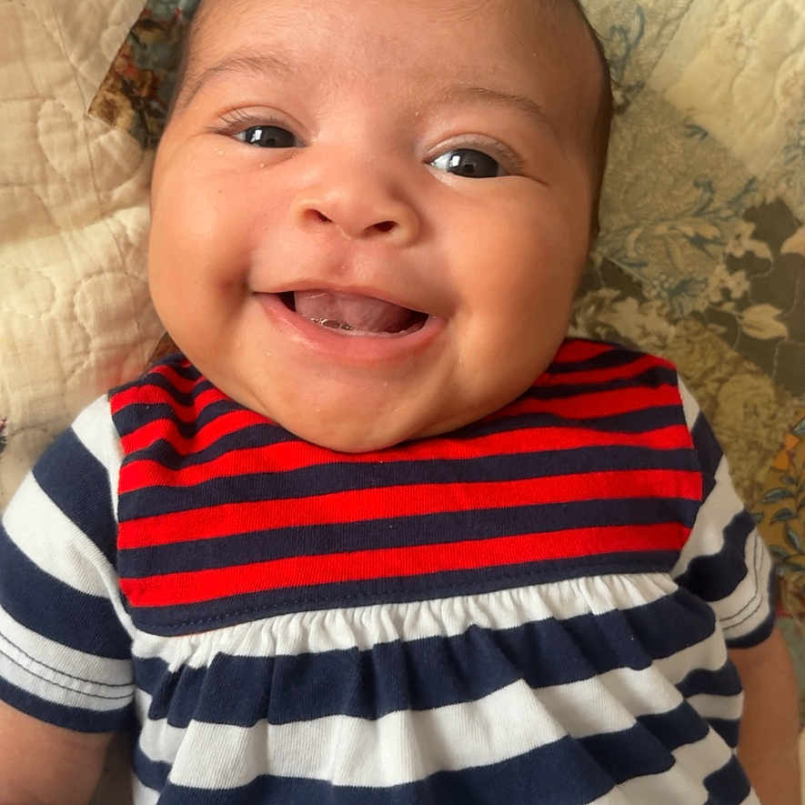 Ilanna is registered to the contest to win money with this photo: baby, smiling, child, infant, striped_clothing, red, white, blue, quilt, patterned_blanket, portrait, happy, cute, young, face, head, lying_down, indoors, person, apparel