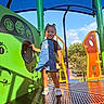 child, toddler, playground, outdoor, smiling, denim_overalls, blue_sky, play_equipment, colorful, green, orange, yellow, purple, trees, sunlight, happy, person, feet, socks, daytime
