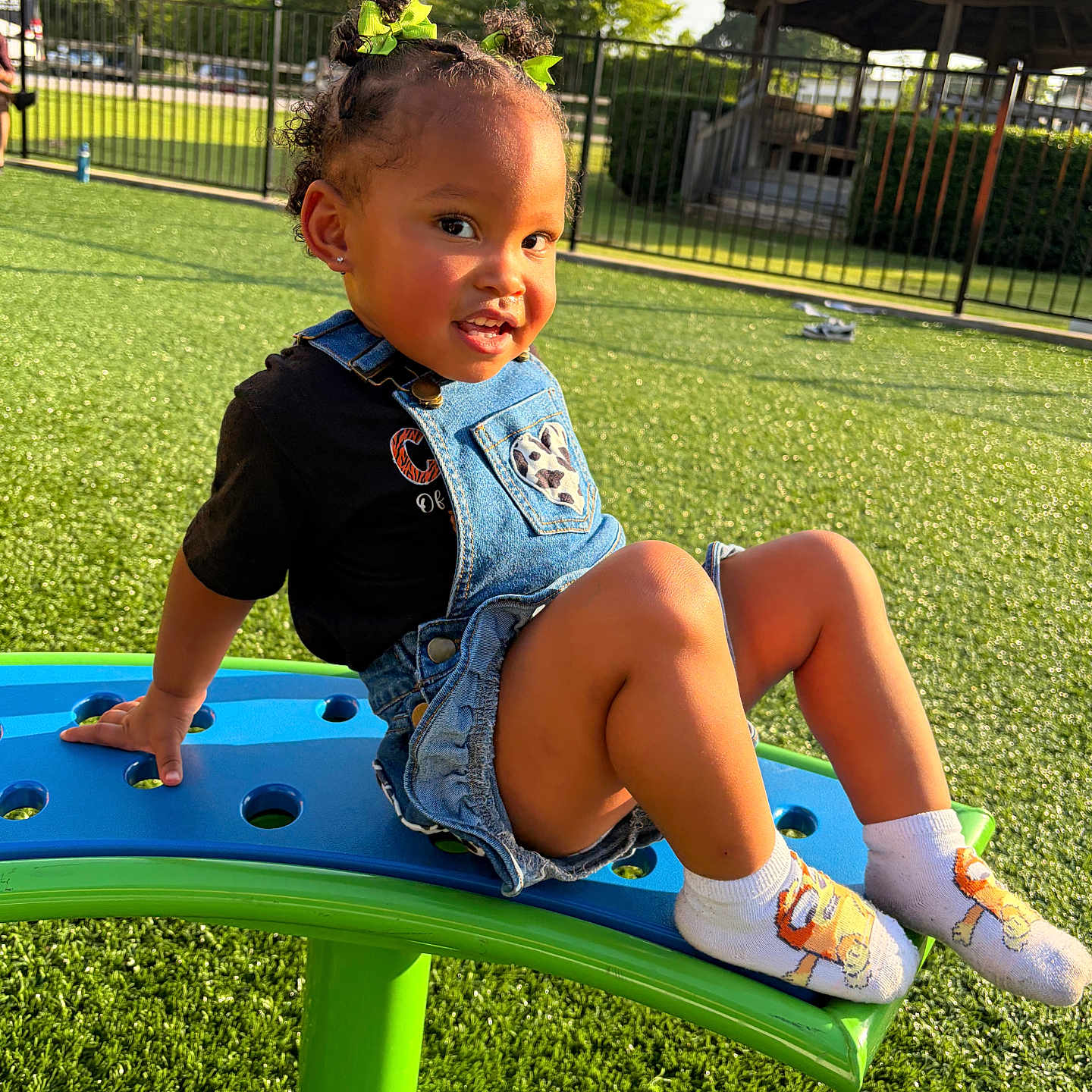 Nayana is registered to the contest to win money with this photo: child, toddler, smiling, playground, outdoor, grass, fence, gazebo, sunlight, hair_bows, clothing, denim_overalls, socks, shoes, play_equipment, green, blue, happy, person, park