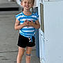 child, smiling, braided_hair, blue_and_white_striped_shirt, black_shorts, pink_crocs, outdoor, concrete, bandage, temporary_tattoos, happy, standing, person, casual_clothing, daylight, trash_bin, metallic_structure, playful, young, kid