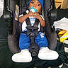 child, toddler, baby, boy, car_seat, pacifier, blue_hat, blue_clothing, white_shoes, sneakers, sitting, strapped_in, wide_eyes, portrait, indoor, vehicle, car_interior, seat_belt, household_items, checkered_blanket