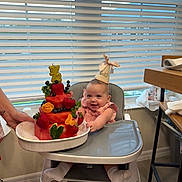 Selena is registered to the contest to win money with this photo: baby, high_chair, party_hat, cake, fruit, strawberry, blueberry, watermelon, orange_slice, celebration, smiling, indoor, kitchen, window_blinds, person, hand, sock, table, floor, happy
