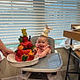 baby, high_chair, party_hat, cake, fruit, strawberry, blueberry, watermelon, orange_slice, celebration, smiling, indoor, kitchen, window_blinds, person, hand, sock, table, floor, happy