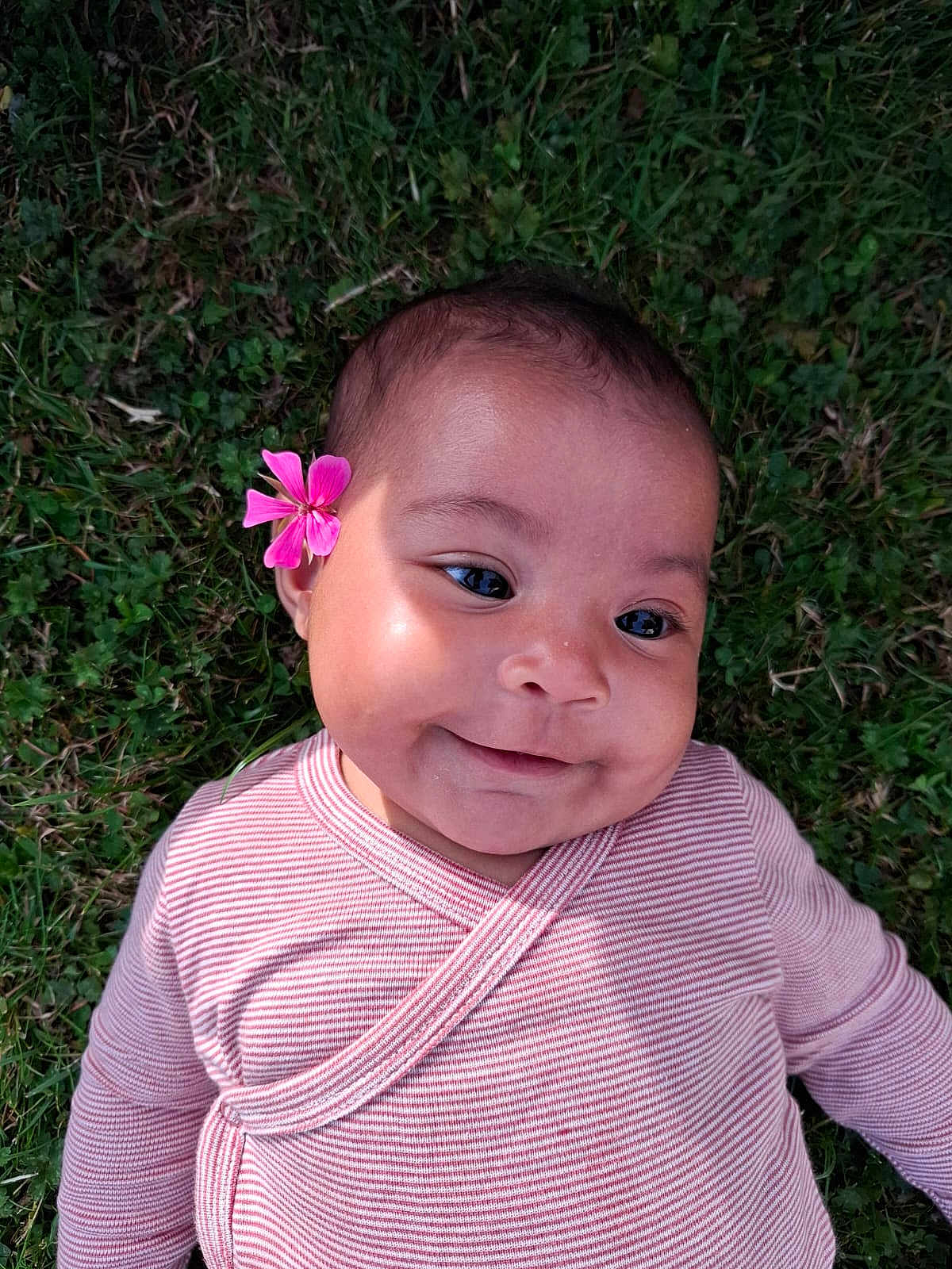 Milaya a rejoint le concours — aidez-le/la à gagner de superbes lots ! baby, child, smiling, flower, pink, striped_clothing, grass, outdoor, cute, face, portrait, head, skin, happy, young_child, infant, nature, laying_down, person, closeup