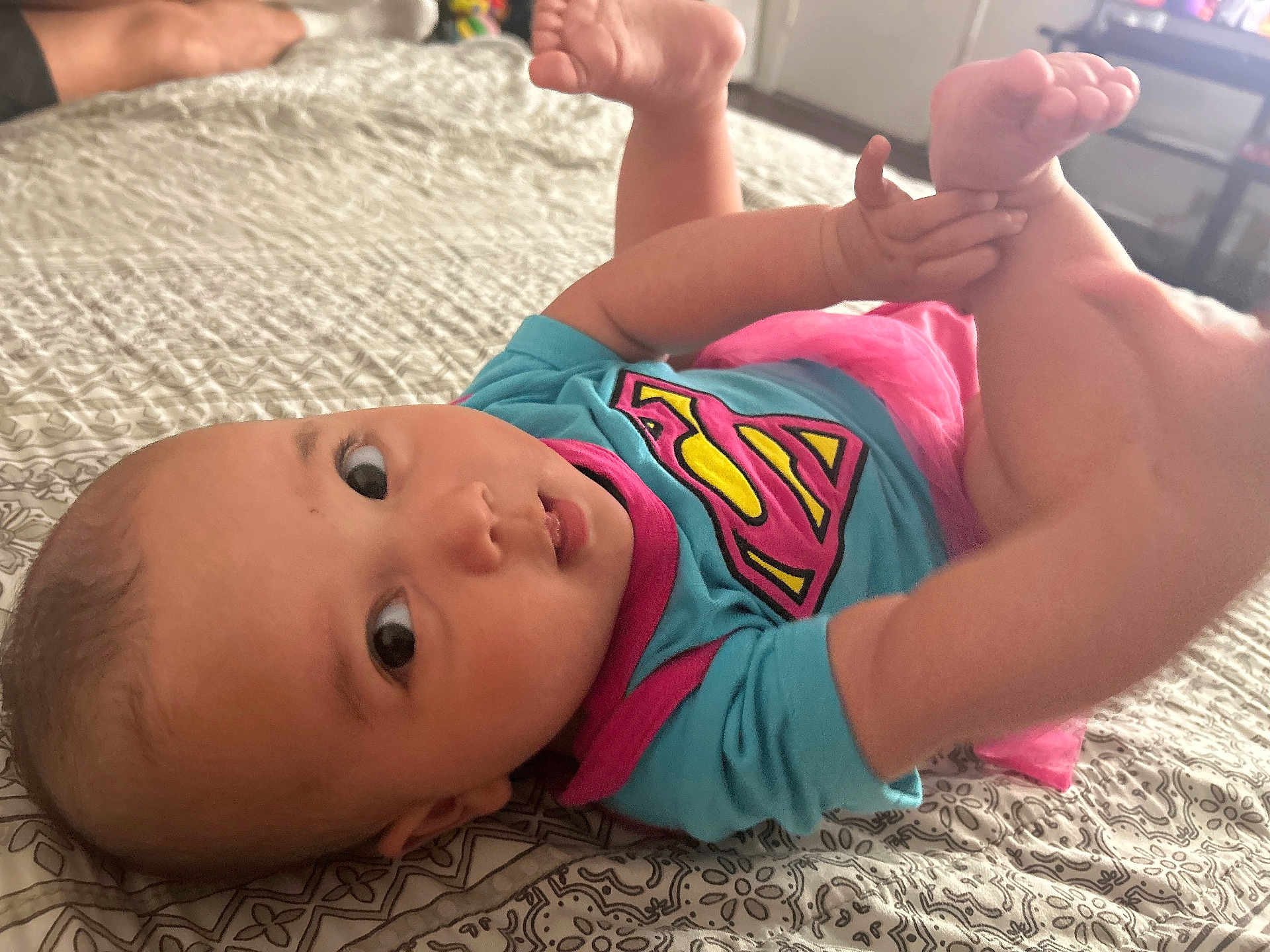 Meyli joined the competition — help win amazing prizes! baby, infant, onesie, superhero_logo, pink_tutu, bed, blanket, patterned_sheet, feet, hands, big_eyes, portrait, close_up, lying_down, skin, cute, indoor, furniture, television, parent_hand