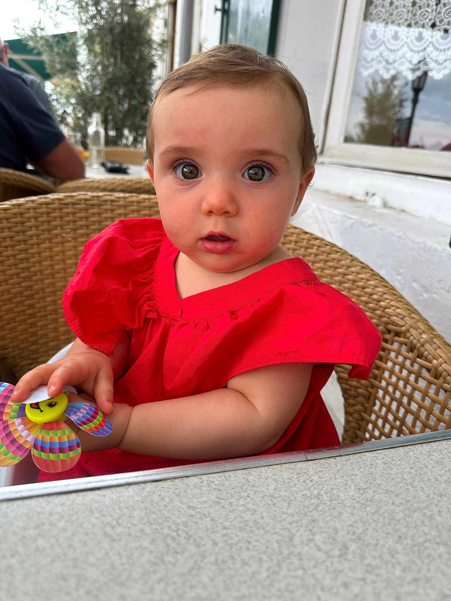 Garance a rejoint le concours — aidez-le/la à gagner de superbes lots ! baby, child, red_dress, wicker_chair, toy, pinwheel, curious, outdoor, cafe, table, person, portrait, cute, innocent, seated, face, expression, colorful, hands, young
