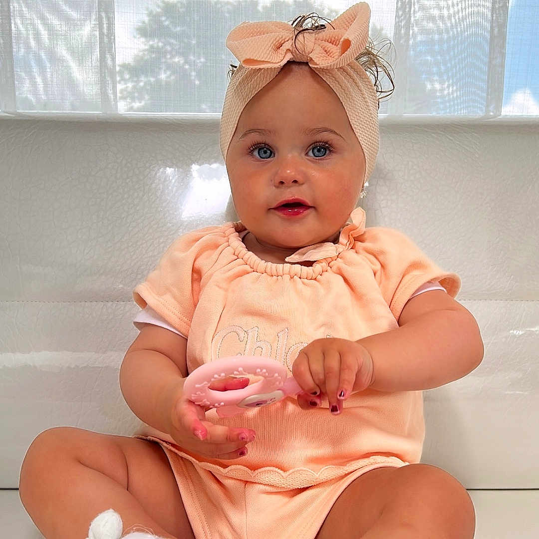 Iris a rejoint le concours — aidez-le/la à gagner de superbes lots ! baby, blue_eyes, bow, child, curious, cute, fingernails, girl, headband, indoor, infant, natural_light, pink_clothing, portrait, sitting, smiling, soft_lighting, toy, white_couch, young_child