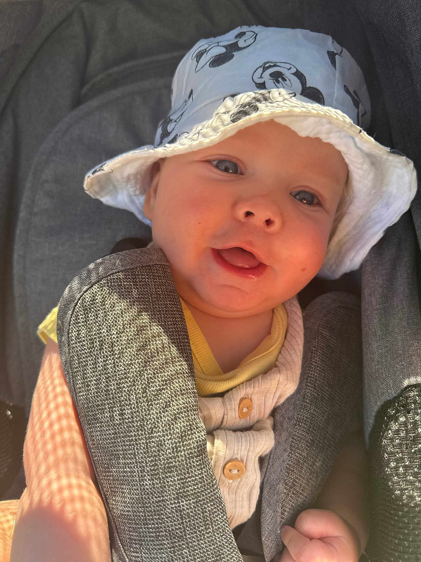 Tobias participe au concours pour gagner de l'argent avec cette photo : baby, child, hat, mickey_mouse, car_seat, smiling, sunlight, face, clothing, seatbelt, infant, cute, portrait, closeup, yellow, white, fabric, skin, hand, outdoor