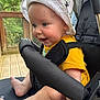 baby, child, stroller, hat, yellow_shirt, smiling, happy, face, blue_eyes, barefoot, outdoor, wooden_deck, greenery, seatbelt, person, infant, cute, sunhat, expression, closeup