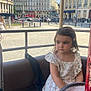 Emma participe au concours pour gagner de l'argent avec cette photo : background, building, carousel, child, city, daylight, floral_dress, girl, hair_clip, outdoor, person, public_space, quiet, seat, side_view, street, sunny, table, thoughtful, urban