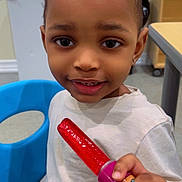 Earl is registered to the contest to win money with this photo: child, smiling, popsicle, red_popsicle, blue_chair, indoor, table, white_shirt, cute, person, face, hand, food, snack, young_child, happy, portrait, sitting, room, furniture