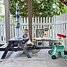 toddler, child, play, toy, picnic_bench, outdoor, patio, fence, kitchen_set, smile, happy, greenery, umbrella, concrete_floor, plastic_toys, casual_clothing, daylight, person, fun, recreation