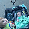 baby, blanket, camouflage, car_seat, child, daytime, family, green_blanket, happy, holding, outdoor, pavement, person, sibling, sidewalk, smile, street, toddler, urban, wagon