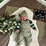 Reginald joined the competition — help win amazing prizes! baby, smiling, knitted_clothing, green_clothing, hat, white_rug, flowers, daisies, wooden_floor, letter_board, text_board, indoor, cozy, cute, baby_accessory, child, happy, laying_down, decor, plant