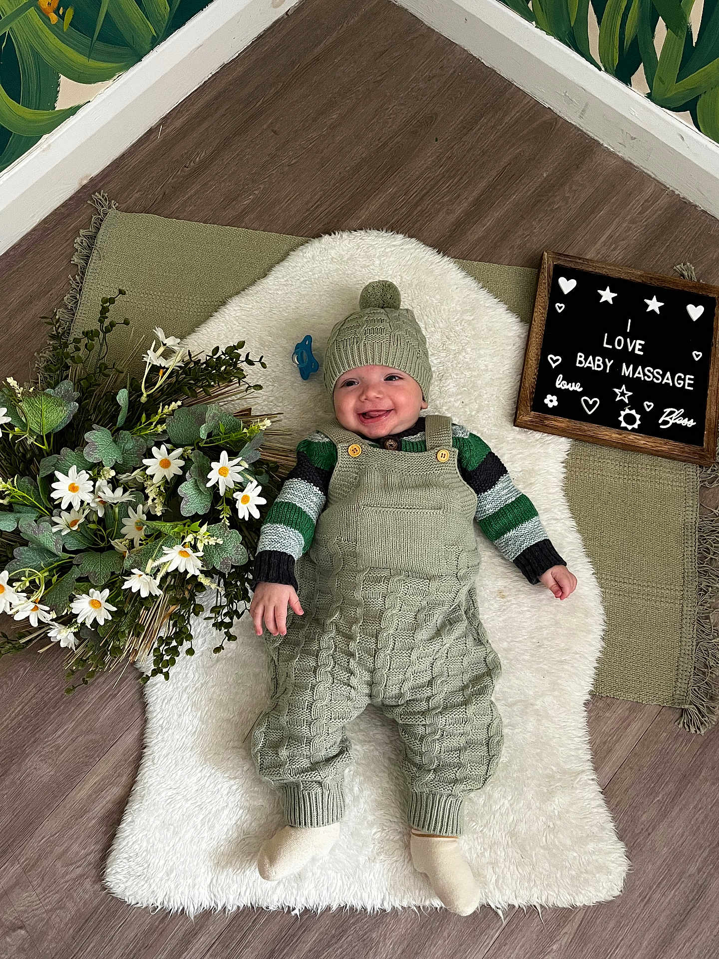 Reginald joined the competition — help win amazing prizes! baby, smiling, knitted_clothing, green_clothing, hat, white_rug, flowers, daisies, wooden_floor, letter_board, text_board, indoor, cozy, cute, baby_accessory, child, happy, laying_down, decor, plant