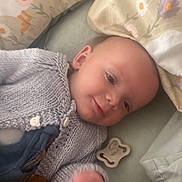 Reginald joined the competition — help win amazing prizes! baby, infant, knitted_sweater, pillow, bed, toy, pacifier, smile, face, child, blanket, soft_toy, sleepwear, cute, indoor, relaxation, comfort, floral_pattern, cushion, lying_down