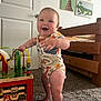 baby, child, smiling, standing, toy, wooden_toy, carpet, bed, furniture, door, animal_paintings, room, indoor, happy, infant, clothing, colorful, play, person, home