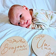 Louka a rejoint le concours — aidez-le/la à gagner de superbes lots ! baby, newborn, infant, milestone_cards, wooden_signs, blanket, bed, pajamas, lying_down, portrait, cute, face, head, eyes, soft_lighting, white_bedding, newborn_photography, wooden_disc, smile, sleepy