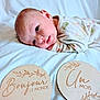 baby, newborn, infant, milestone_cards, wooden_signs, blanket, bed, pajamas, lying_down, portrait, cute, face, head, eyes, soft_lighting, white_bedding, newborn_photography, wooden_disc, smile, sleepy