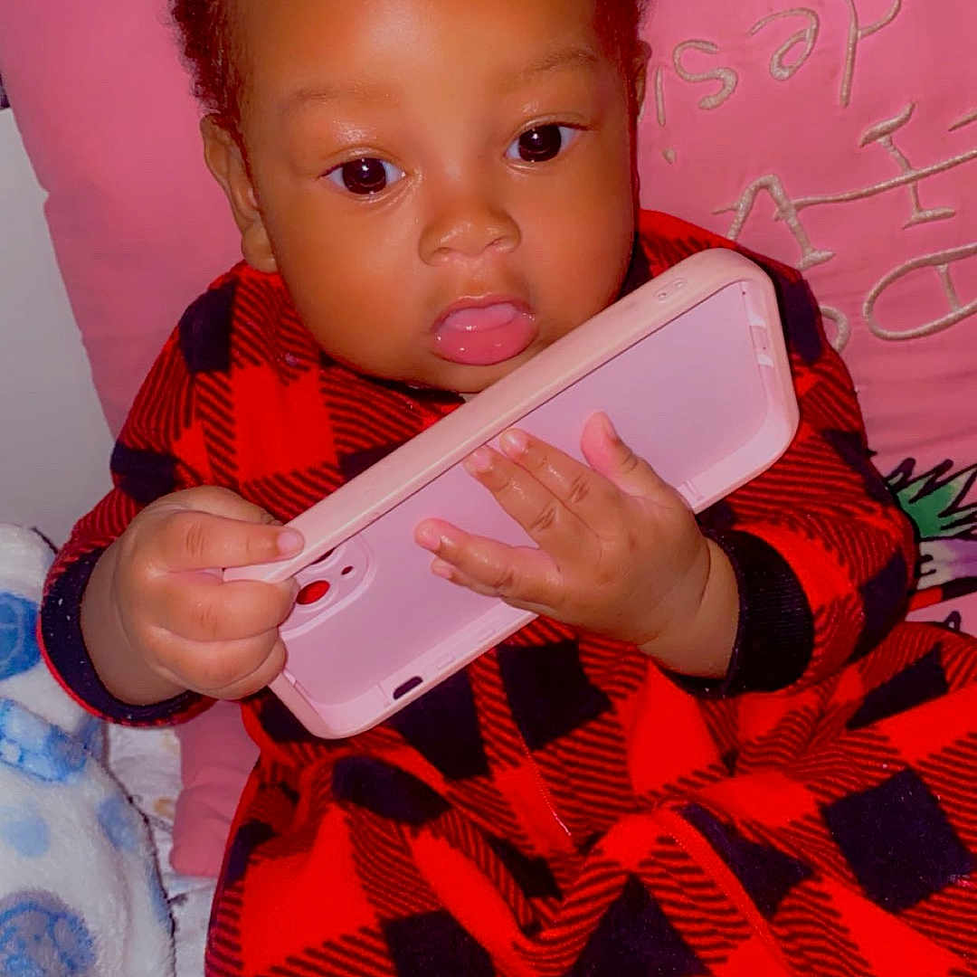 Jeanclaude is registered to the contest to win money with this photo: baby, bed, black, blanket, child, clothing, cozy, curly_hair, dark_skin, eyes, face, indoors, infant, phone, pink, portrait, red, sitting, smartphone, tongue