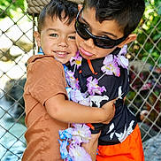 Elijah joined the competition — help win amazing prizes! child, hug, boys, sunglasses, leis, flower_garland, orange_shorts, brown_shirt, plaid_shorts, smile, outdoor, fence, greenery, tropical, happy, friendship, portrait, casual_clothing, playful, summer