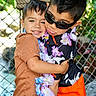 child, hug, boys, sunglasses, leis, flower_garland, orange_shorts, brown_shirt, plaid_shorts, smile, outdoor, fence, greenery, tropical, happy, friendship, portrait, casual_clothing, playful, summer