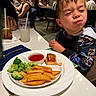 boy, broccoli, child, clothing, dining, drink, expression, face, fish, food, fork, glass, indoor, ketchup, napkin, person, plate, potato_wedges, restaurant, table