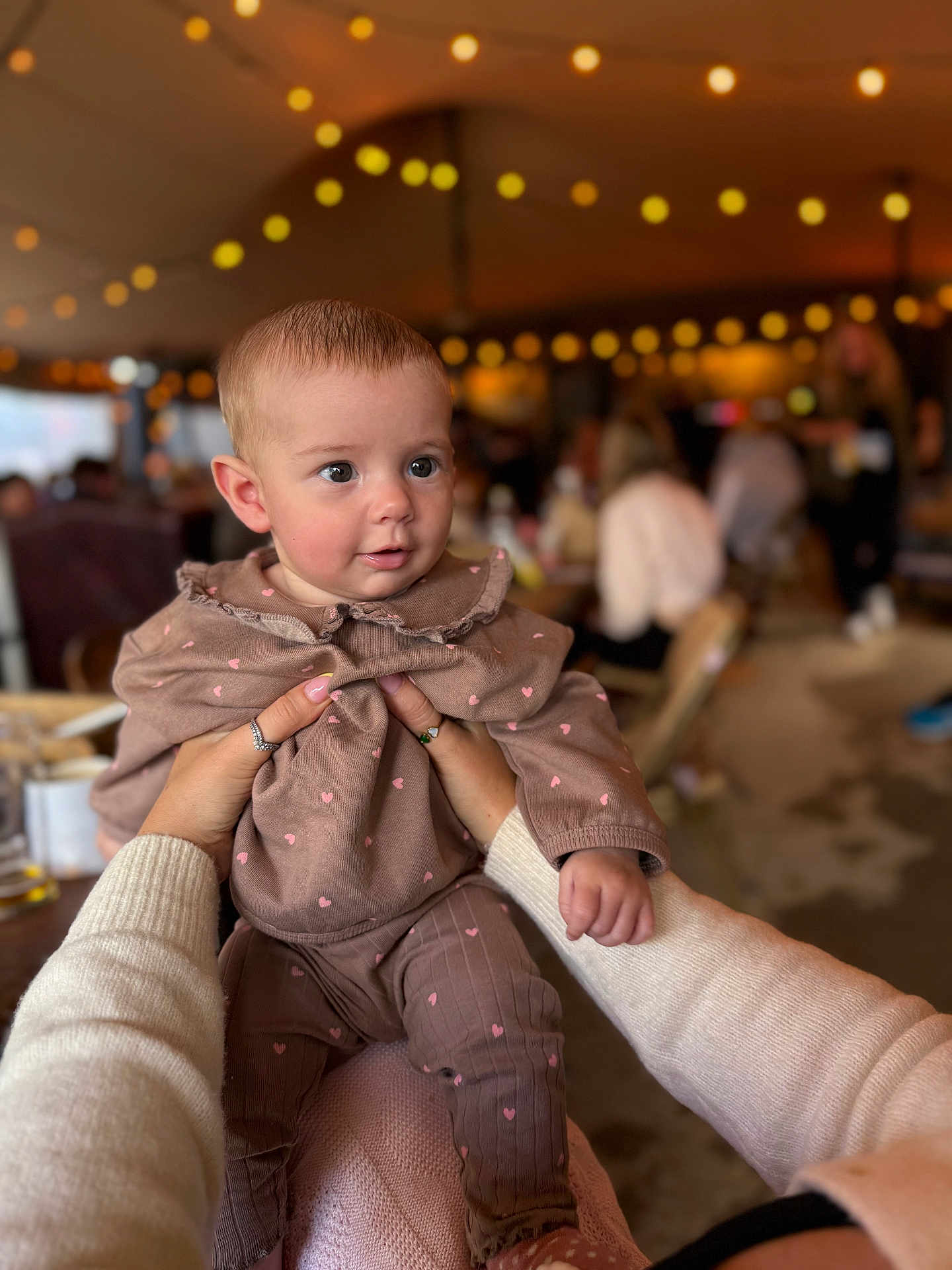 Bobbie is registered to the contest to win money with this photo: baby, child, hands, clothing, heart_pattern, indoor, string_lights, blurred_background, people, warm_lighting, cute, portrait, person, infant, sweater, pink, brown, holding, face, expression