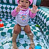 toddler, child, inflatable_pool, water, splashing, pink_hat, swimsuit, toys, colorful, outdoor, summer, playful, happy, sitting, water_toys, backyard, sunlight, patterned_pool, curious, baby