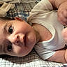 baby, infant, child, lying_down, wide_eyes, looking_at_camera, face, smiling, white_clothing, onesie, blanket, plaid, bed, cozy, indoor, portrait, close_up, hands, bedding, soft_lighting