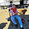 elderly_man, baby, dog, camping, outdoor, chair, camper, grass, sunlight, pet, person, jeans, sneakers, hat, glasses, folding_chair, red_shirt, golden_retriever, family, nature