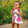 barefoot, blue, bows, child, cute, dress, flowers, garden, greenery, happy, nature, outdoor, portrait, red, smiling, standing, sunlight, toddler, white, yellow_flowers