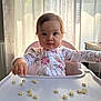 baby, child, high_chair, snack, food, pajamas, floral_pattern, table, indoor, curtains, window, light, face, person, cute, expression, tabletop, feeding, infant, seat