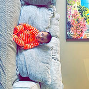 Dilpreet joined the competition — help win amazing prizes! baby, infant, orange_clothing, striped_onesie, couch, pillows, living_room, painting, lamp, indoor, seated, child, furniture, home, cozy, curious, soft_texture, colorful_art, relaxed, portrait