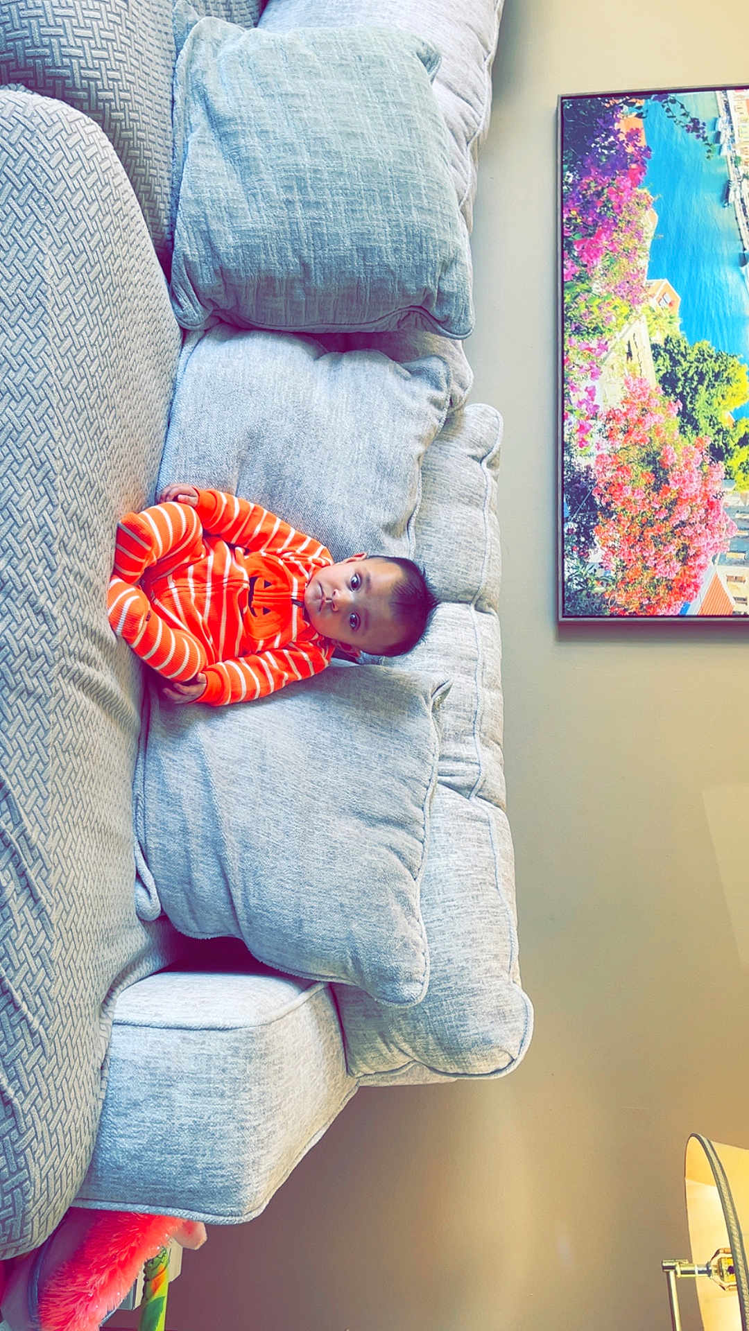 Dilpreet joined the competition — help win amazing prizes! baby, infant, orange_clothing, striped_onesie, couch, pillows, living_room, painting, lamp, indoor, seated, child, furniture, home, cozy, curious, soft_texture, colorful_art, relaxed, portrait