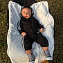 baby, infant, blanket, outdoor, grass, balloon, footwear, black_clothing, smiling, face, person, sportswear, jacket, pants, shoes, happy, child, lying_down, celebration, soft_texture