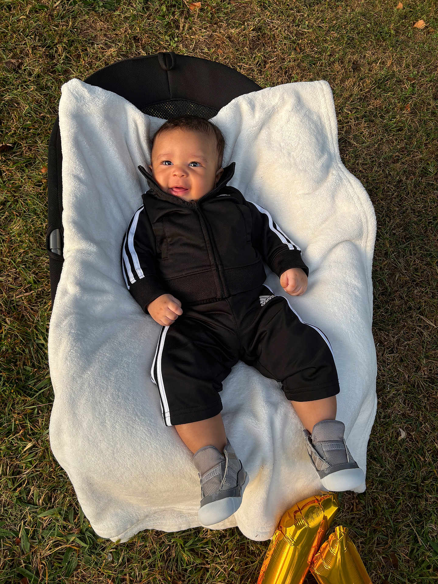 Anthony joined the competition — help win amazing prizes! baby, infant, blanket, outdoor, grass, balloon, footwear, black_clothing, smiling, face, person, sportswear, jacket, pants, shoes, happy, child, lying_down, celebration, soft_texture
