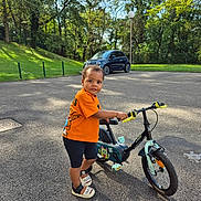 Dream participe au concours pour gagner de l'argent avec cette photo : toddler, child, bicycle, orange_shirt, shorts, outdoor, park, sunny_day, asphalt, car, trees, greenery, footwear, sidewalk, person, play, summer, happy, young, vehicle