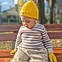 toddler, child, yellow_hat, yellow_gloves, striped_sweater, beige_pants, wooden_bench, outdoor, autumn, sunlight, portrait, knitwear, cute, young_child, seated, focused_expression, daylight, nature_background, casual_clothing, seasonal