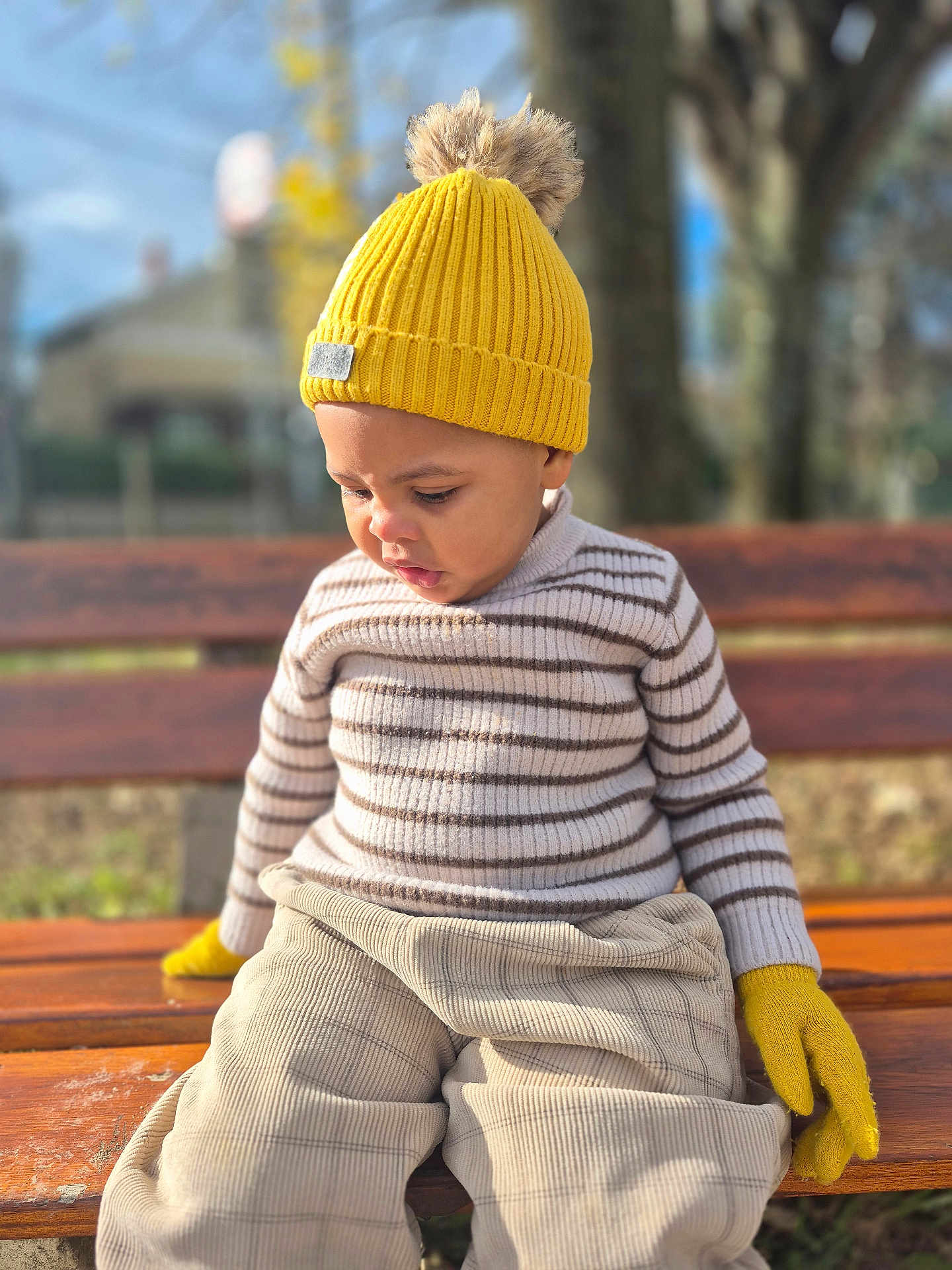 Dream a rejoint le concours — aidez-le/la à gagner de superbes lots ! toddler, child, yellow_hat, yellow_gloves, striped_sweater, beige_pants, wooden_bench, outdoor, autumn, sunlight, portrait, knitwear, cute, young_child, seated, focused_expression, daylight, nature_background, casual_clothing, seasonal