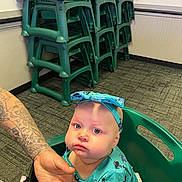 Mielani joined the competition — help win amazing prizes! baby, child, high_chair, tattoo, hand, green_chair, stacked_chairs, headband, blue_clothing, indoor, carpet, wall, person, infant, seated, expression, holding_hand, patterned_floor, casual, portrait