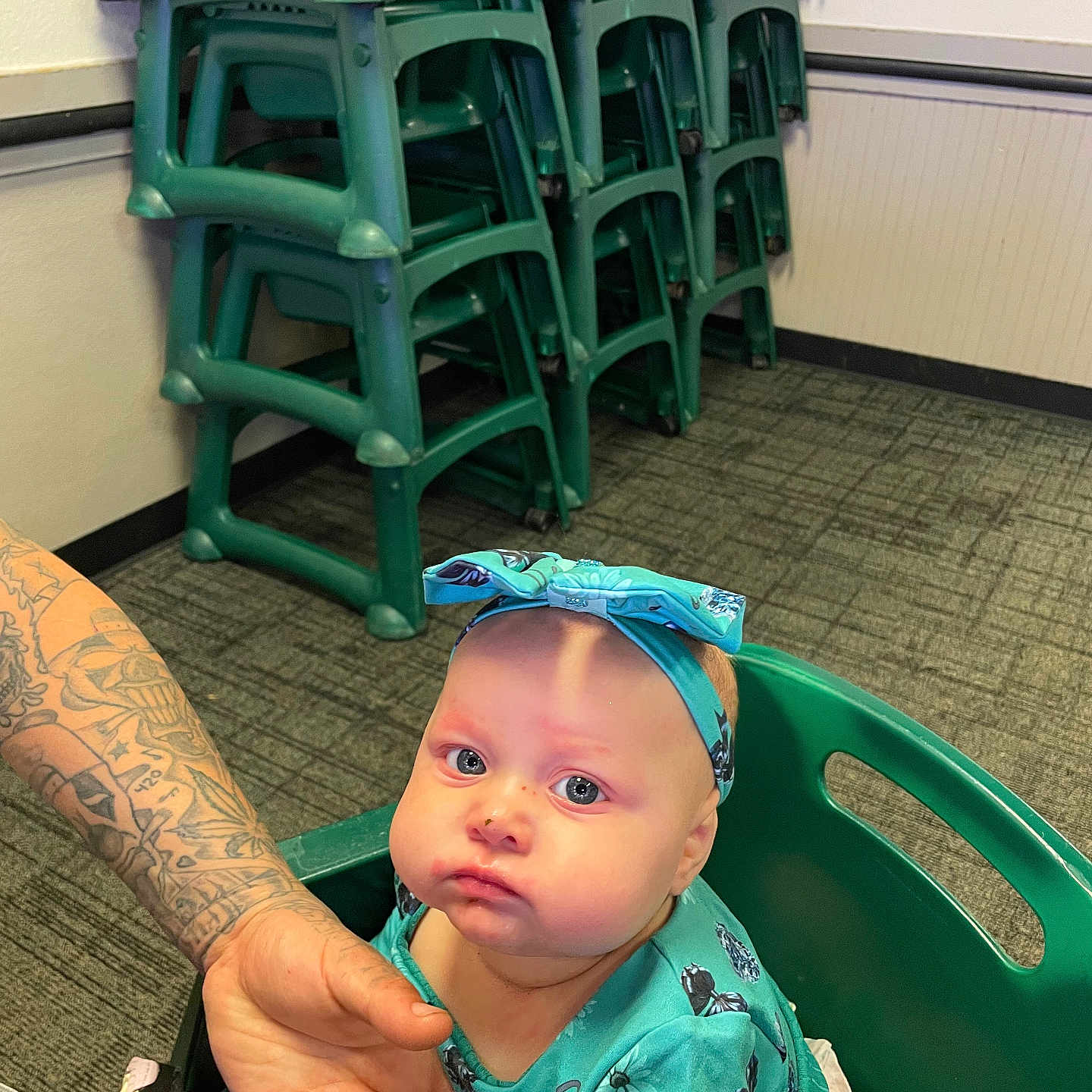 Mielani joined the competition — help win amazing prizes! baby, child, high_chair, tattoo, hand, green_chair, stacked_chairs, headband, blue_clothing, indoor, carpet, wall, person, infant, seated, expression, holding_hand, patterned_floor, casual, portrait