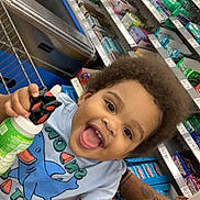 Gianni is registered to the contest to win money with this photo: toddler, child, shopping_cart, grocery_store, smiling, curly_hair, toy, bottle, clothing, shelf, products, colorful, happy, person, retail, shopping, food, store, aisle, casual