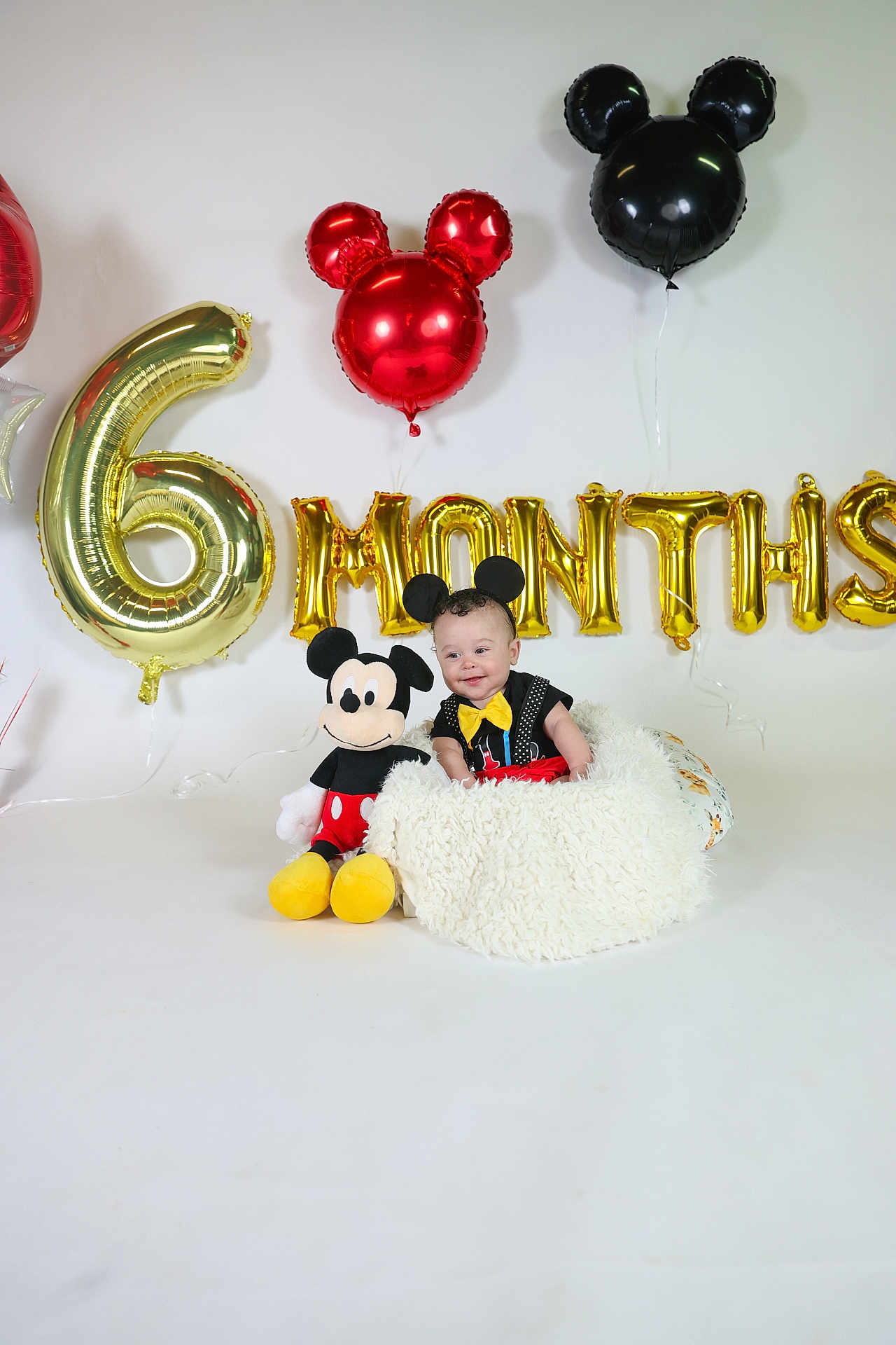 Kilynn is registered to the contest to win money with this photo: baby, child, mickey_mouse, plush_toy, balloons, celebration, birthday, party, costume, blanket, white_background, smiling, yellow_bowtie, black_clothing, red_shorts, indoor, decorations, festive, headband, cute