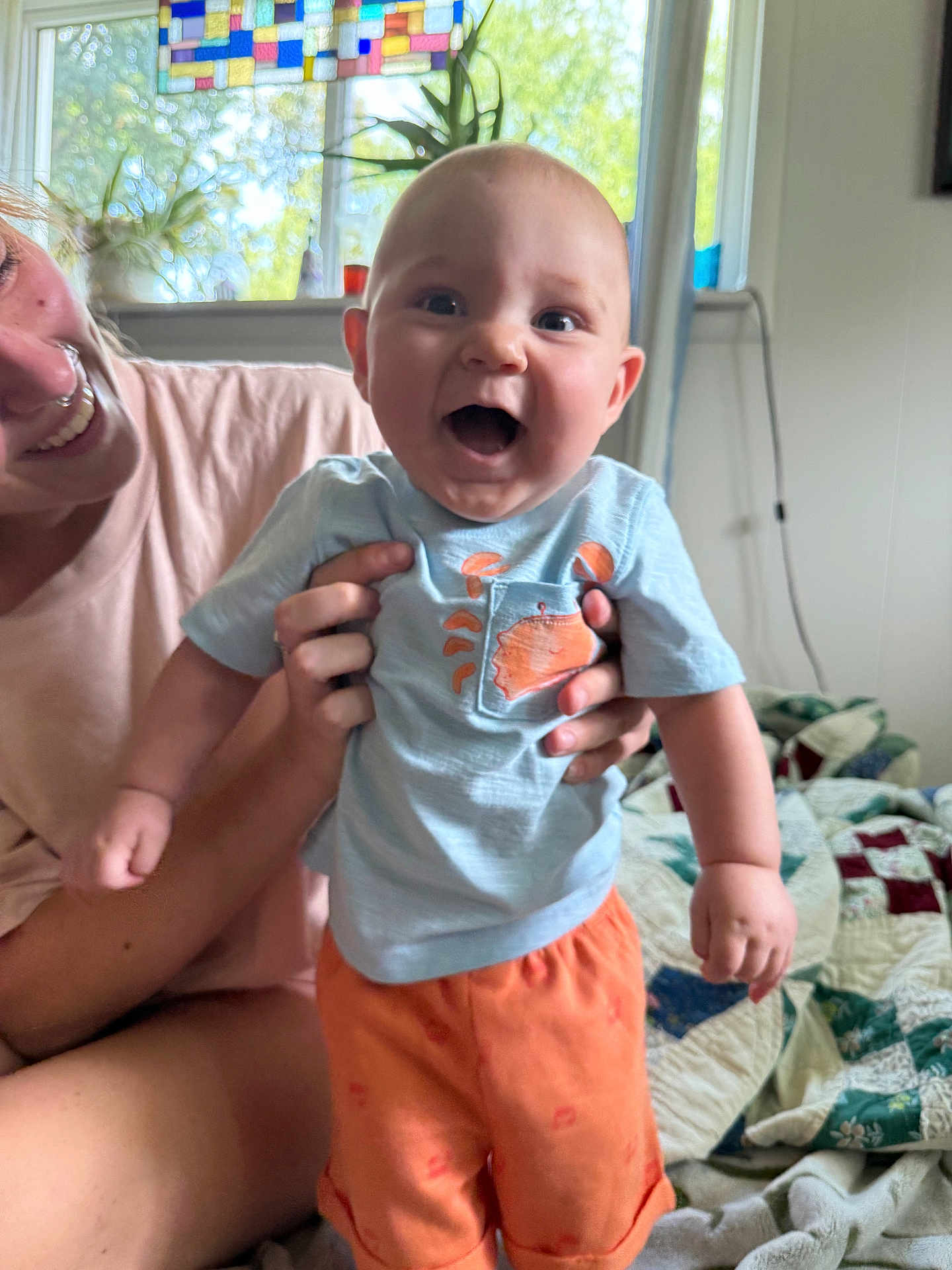 Faust joined the competition — help win amazing prizes! baby, child, smiling, happy, person, indoor, quilts, window, stained_glass, clothing, orange_shorts, blue_shirt, adult, hand, face, room, bed, plant, light, casual