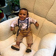 Kyan is registered to the contest to win money with this photo: baby, toddler, child, smiling, sitting, indoor, chair, plush, clothing, bow_tie, suspenders, pants, shoes, happy, face, person, plant, pot, home, cozy
