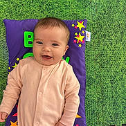 Harriet is registered to the contest to win money with this photo: baby, smiling, infant, onesie, pillow, purple, stars, green_mat, happy, child, cute, playmat, indoors, face, portrait, young_child, lying_down, colorful, fabric, soft_toys